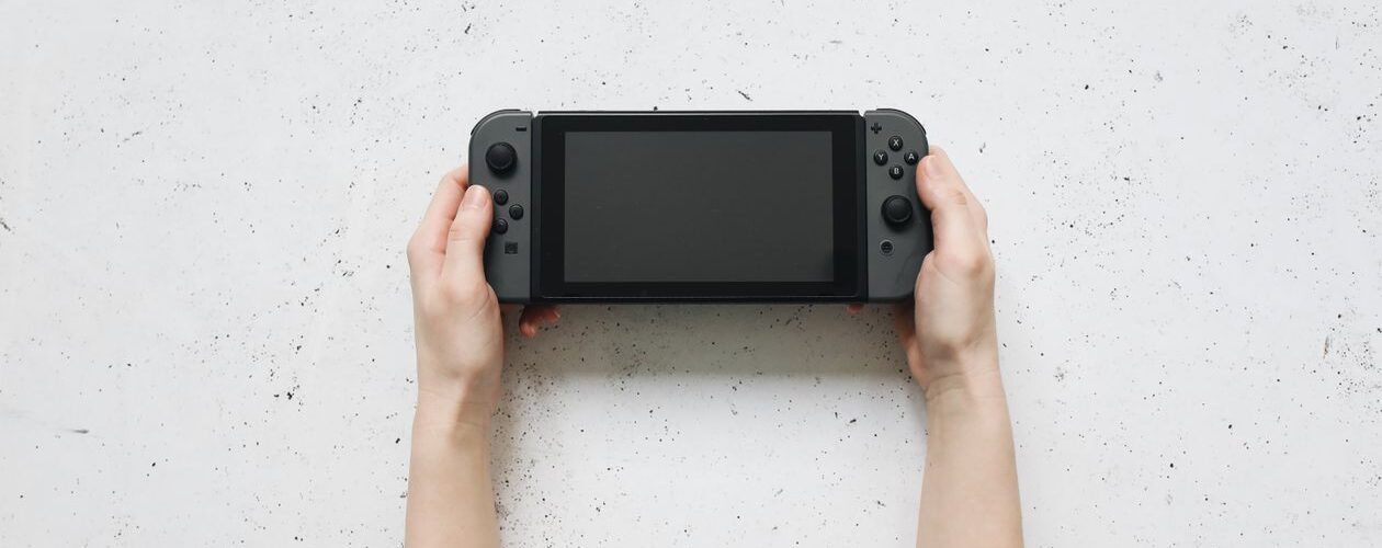 Two hands holding a gray handheld game console with a blank screen against a white speckled wall